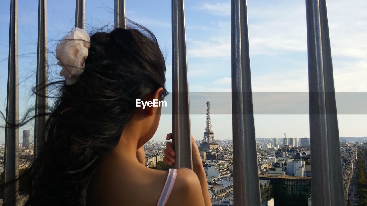 Rear view of girl looking at eiffel tower in city seen through fence