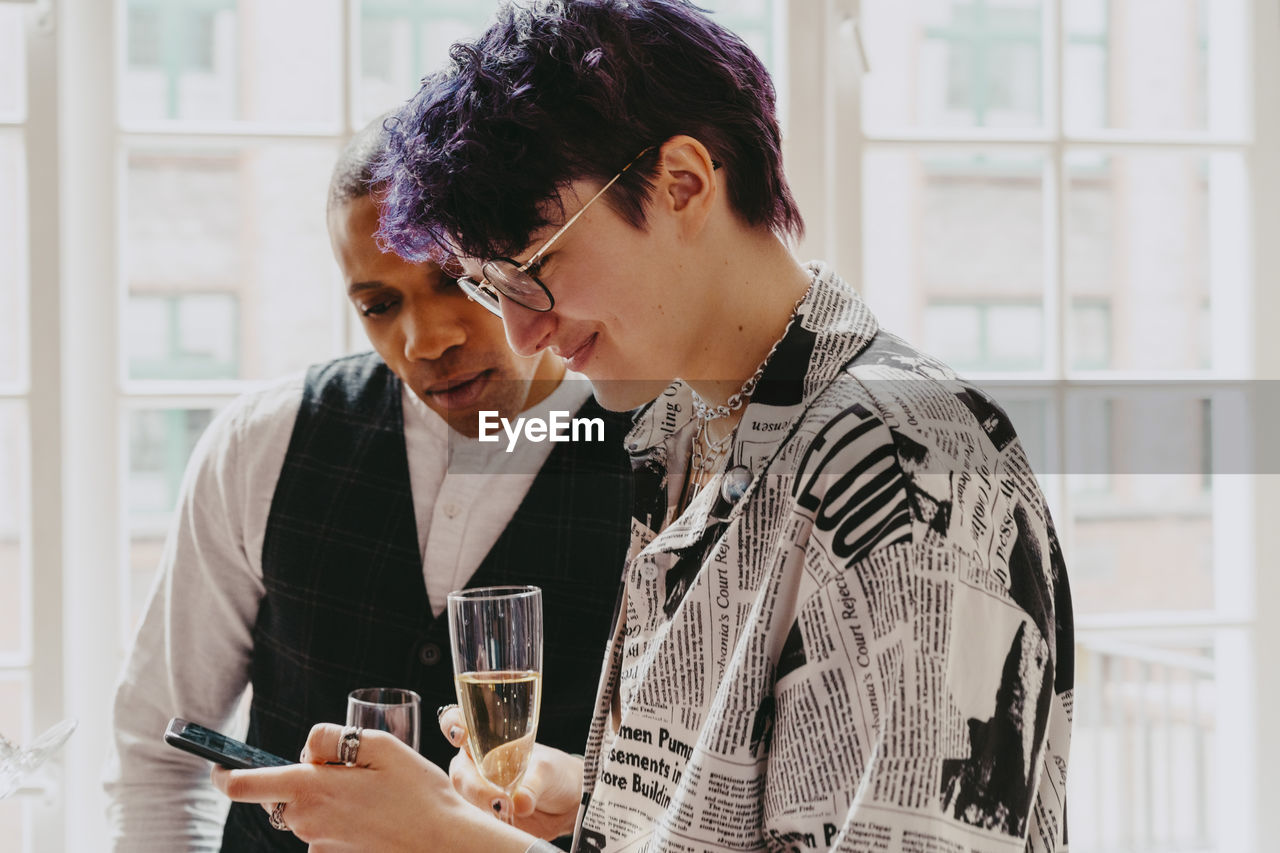 Smiling non-binary person sharing smart phone with male colleague during event at convention center