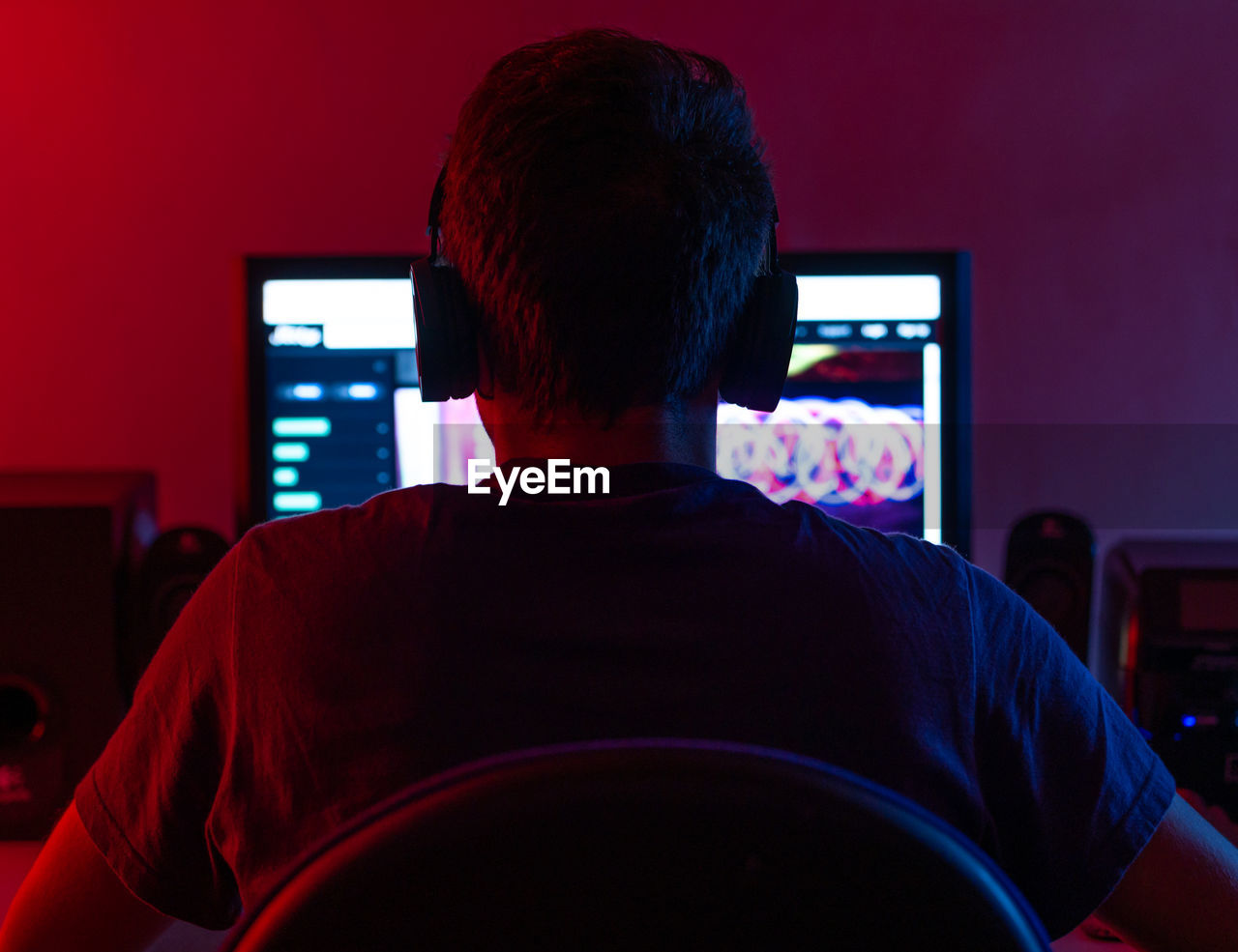 Back view of calm young brunet in casual wear and wireless headset browsing contemporary computer with blank screen while sitting at table in dark room