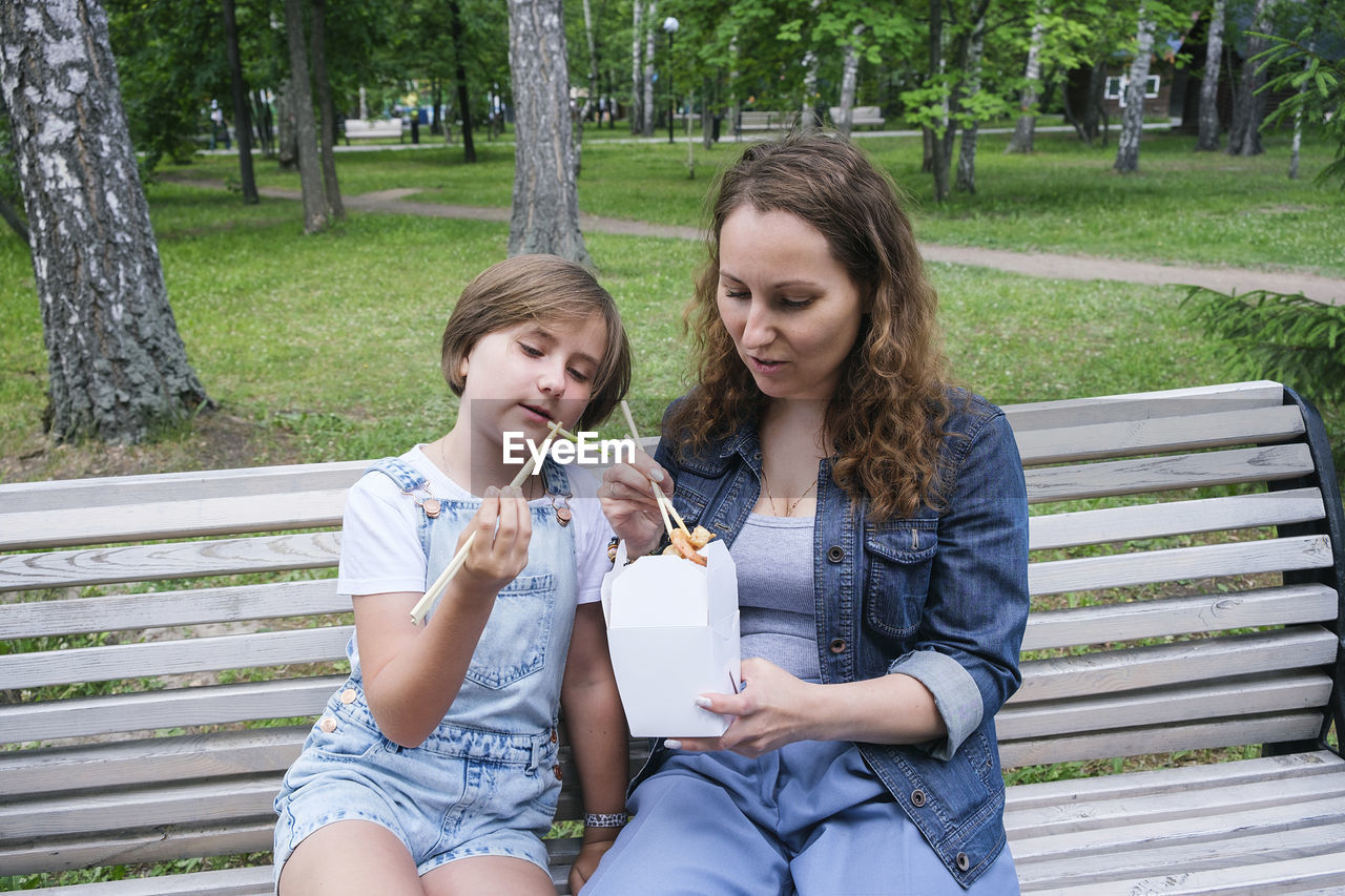 Mid adult mature woman and teenage girl on a summer day in the park has lunch with noodles wok