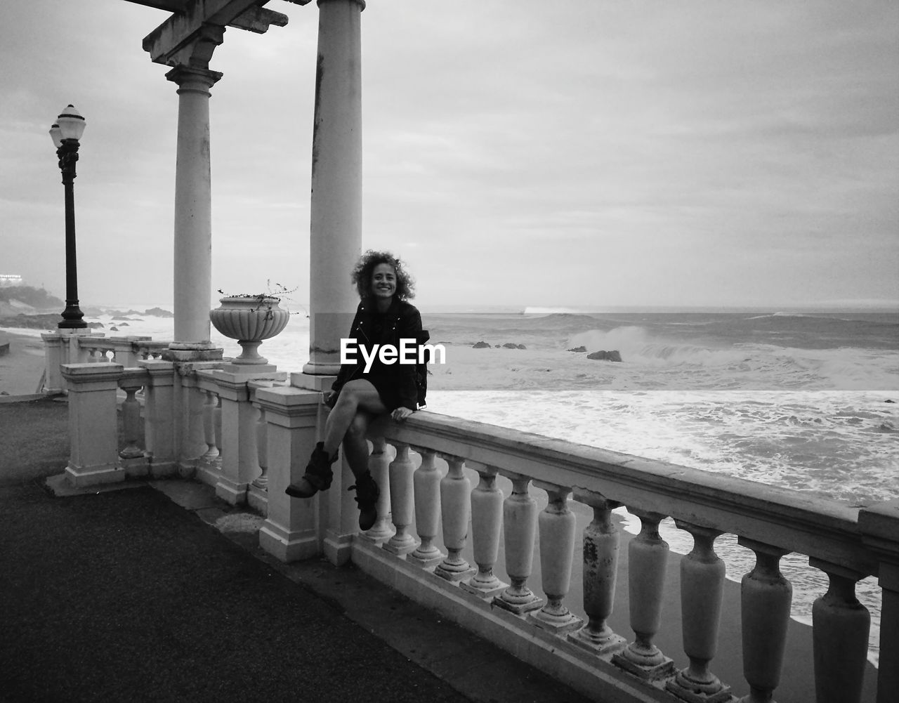 Portrait of woman sitting on railing by sea against sky