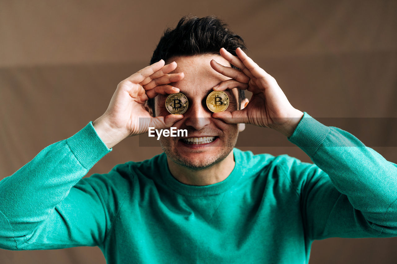 Cheerful male broker in green wear covering eyes with metal crypto coins on brown background