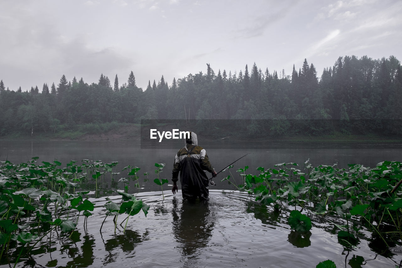 Rear view of man holding fishing rod while standing in lake against trees and sky during foggy weather