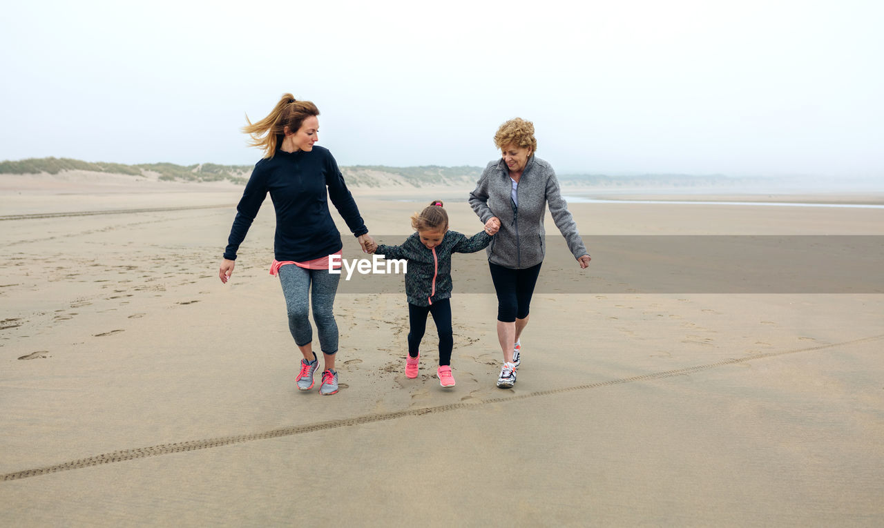 Playful woman walking with mother and daughter at beach against sky