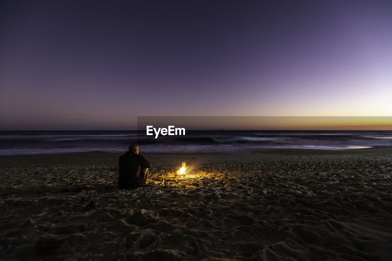 Rear view of a lonely guy and bonfire on the beach at sunset