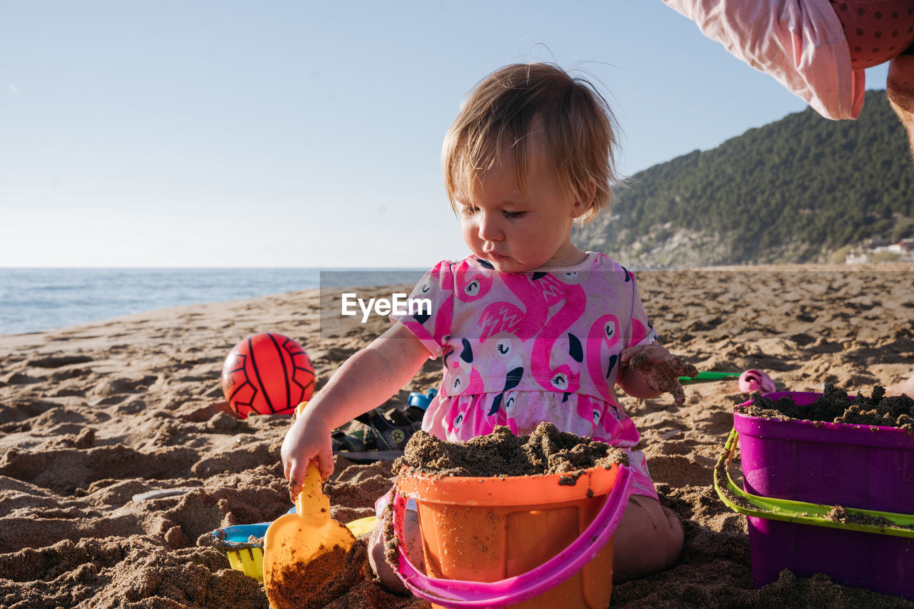 Baby girl in pink dress sitting in a sand with sand toys at the beach