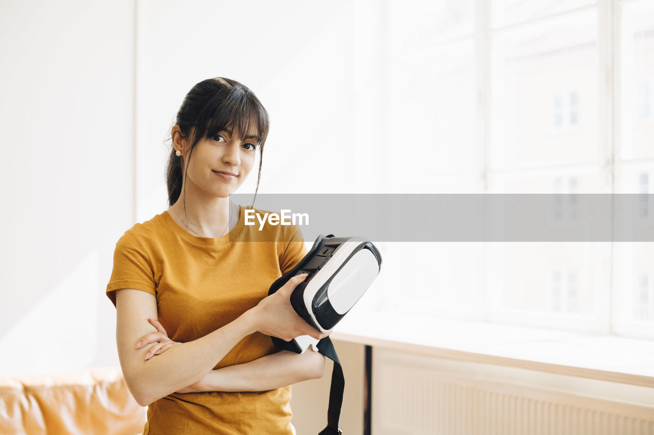 Portrait of female entrepreneur holding virtual reality simulator while standing by window