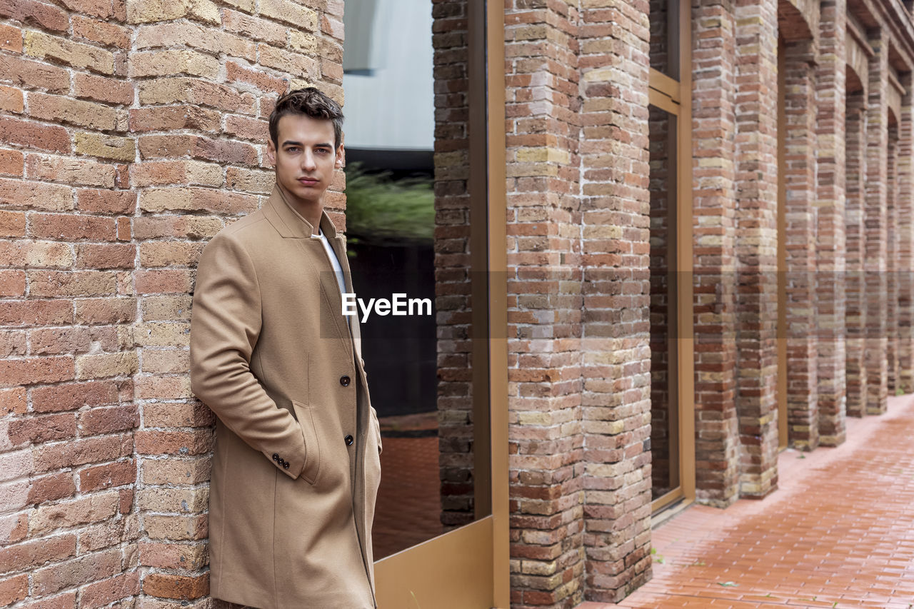 one person, adult, brick, architecture, standing, brick wall, men, young adult, coat, wall, clothing, spring, building exterior, outerwear, business, city, looking, built structure, portrait, three quarter length, day, hands in pockets, textile, lifestyles, businessman, outdoors, jacket, wall - building feature, fashion, formal wear, looking at camera, business finance and industry