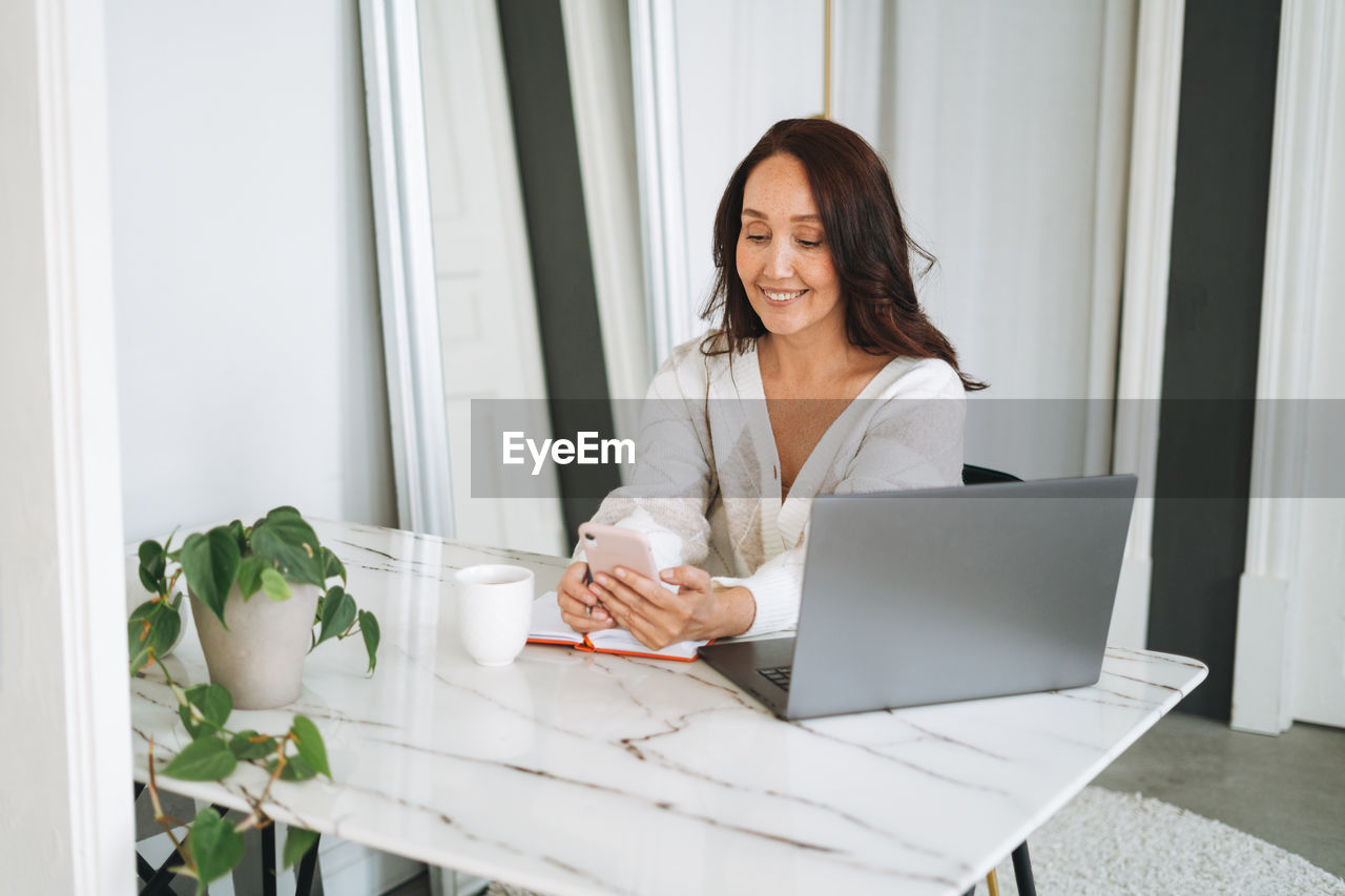 Smiling woman with long hair in white cardigan working on laptop using mobile phone in office