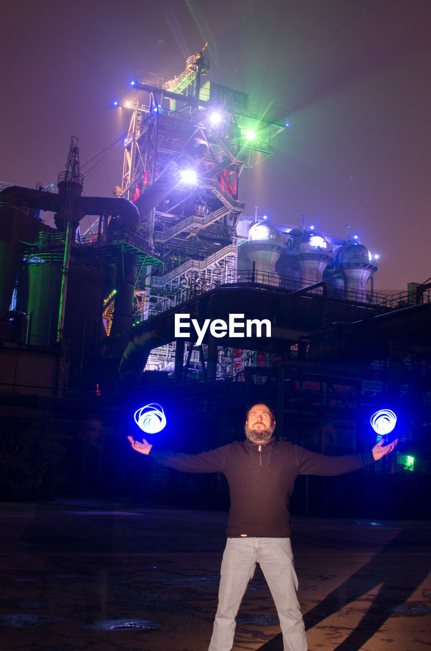 Man standing against illuminated factory against sky at night