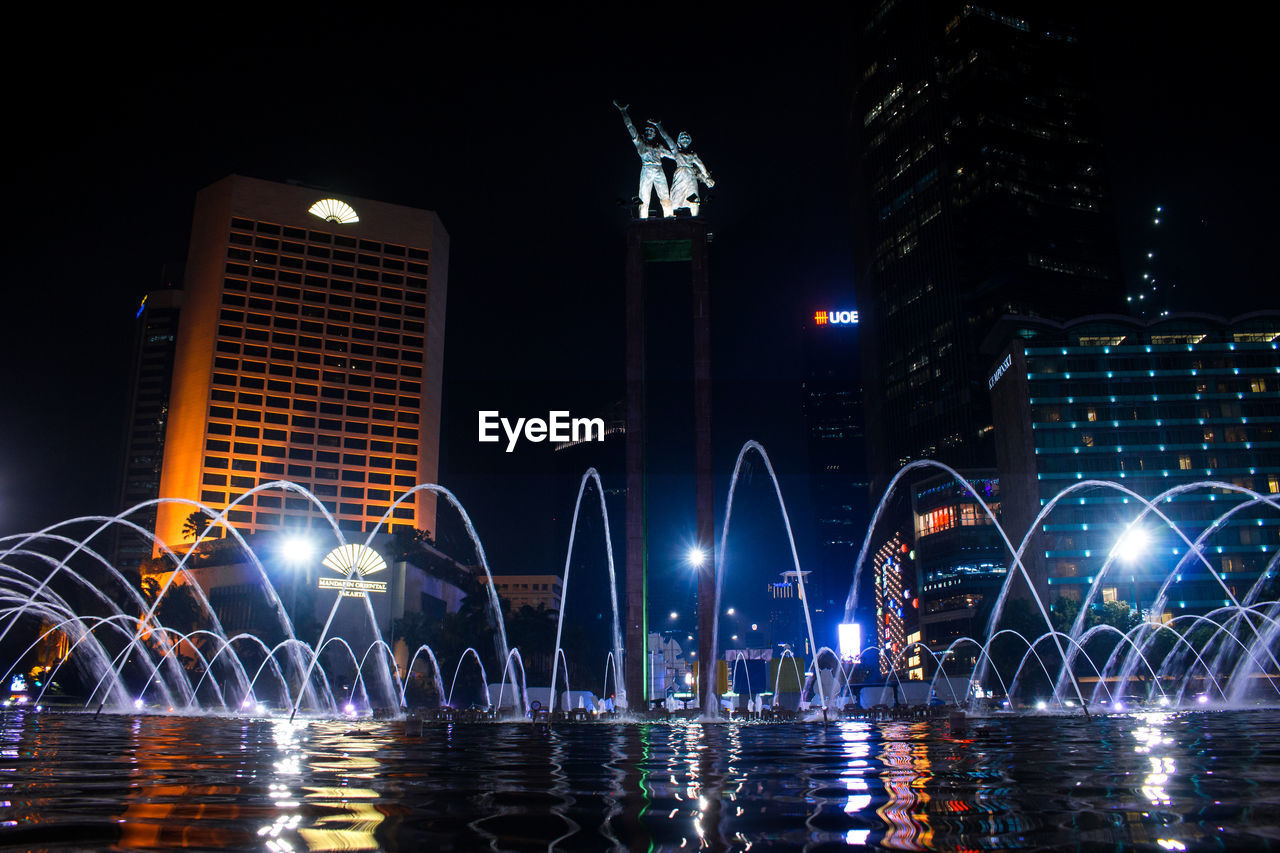 Water fountain against illuminated buildings in city at night