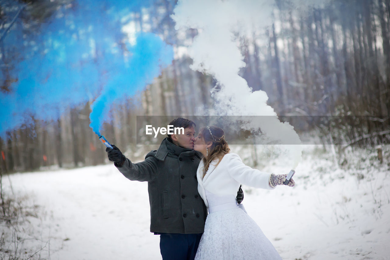 Smiling wedding couple holding distress flares while standing at forest during winter