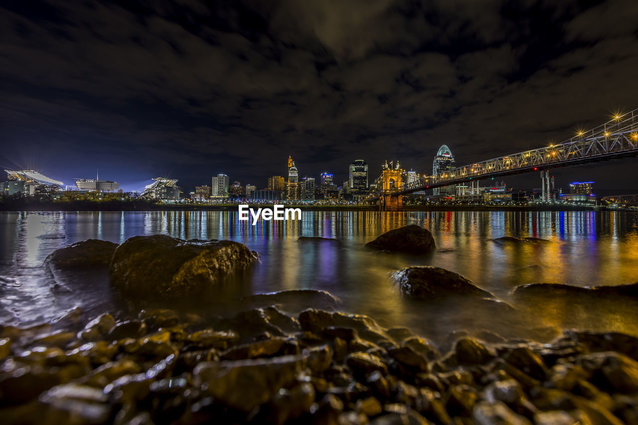 Illuminated bridge and buildings with reflection in river against sky