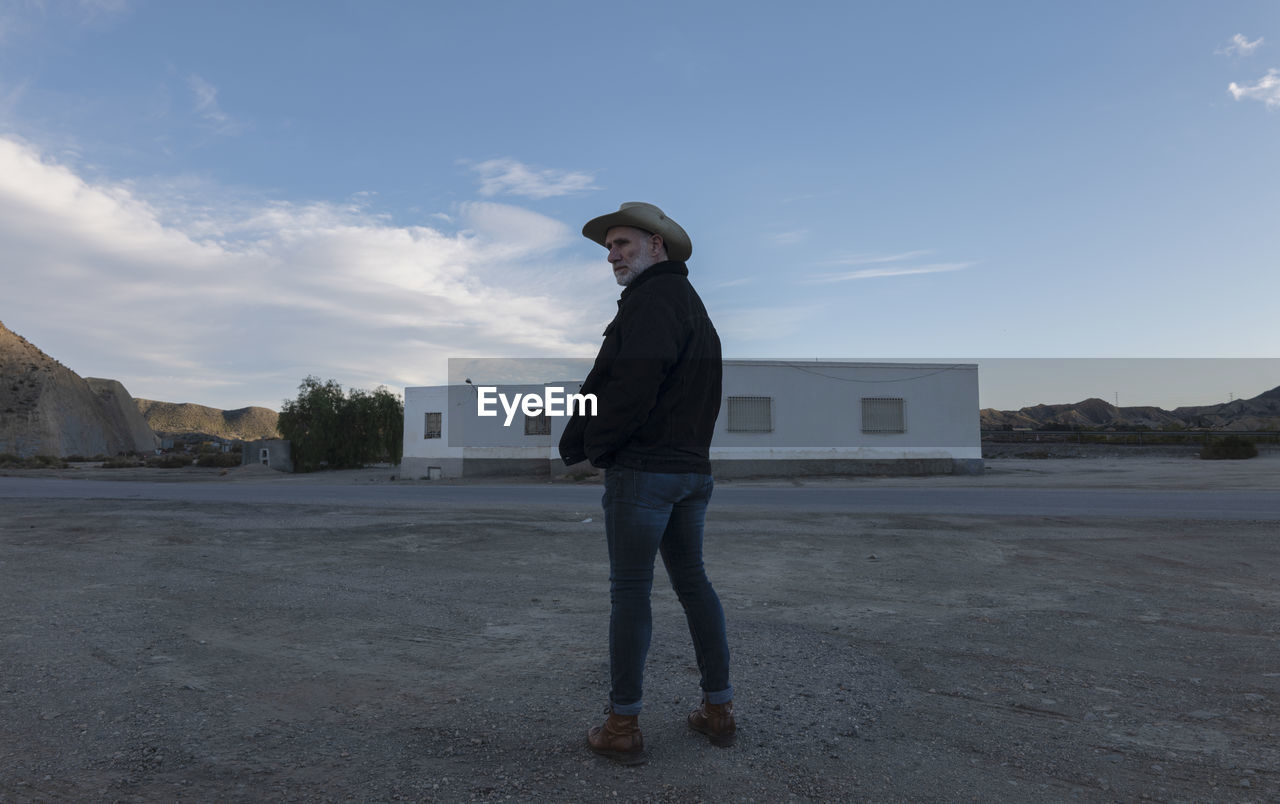 Adult man in cowboy hat standing in front of white building against sky