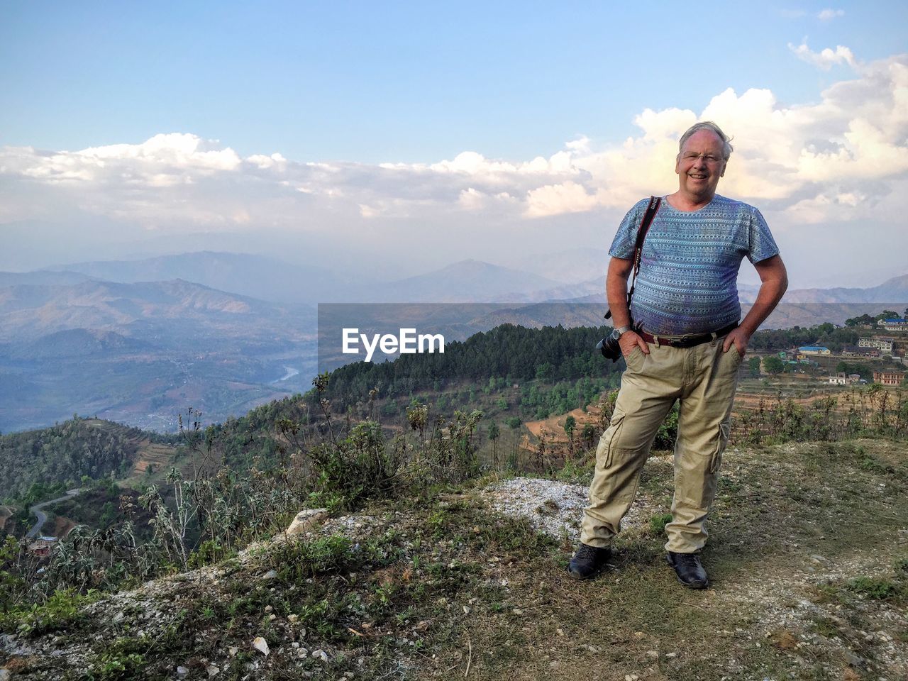 Smiling senior man with hands in pockets standing on cliff against cloudy sky