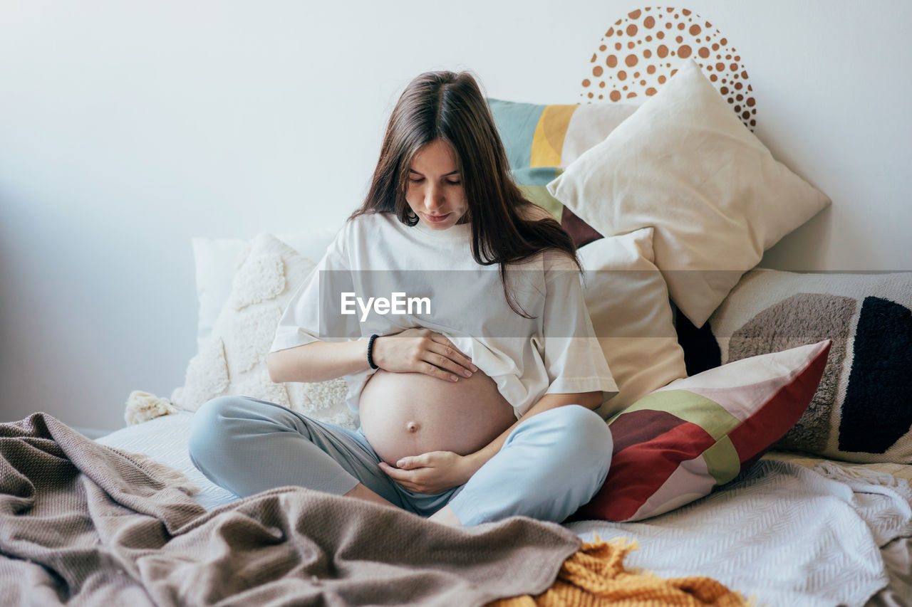 A pregnant woman sitting on the bed in homewear gently holding her pregnant tummy.
