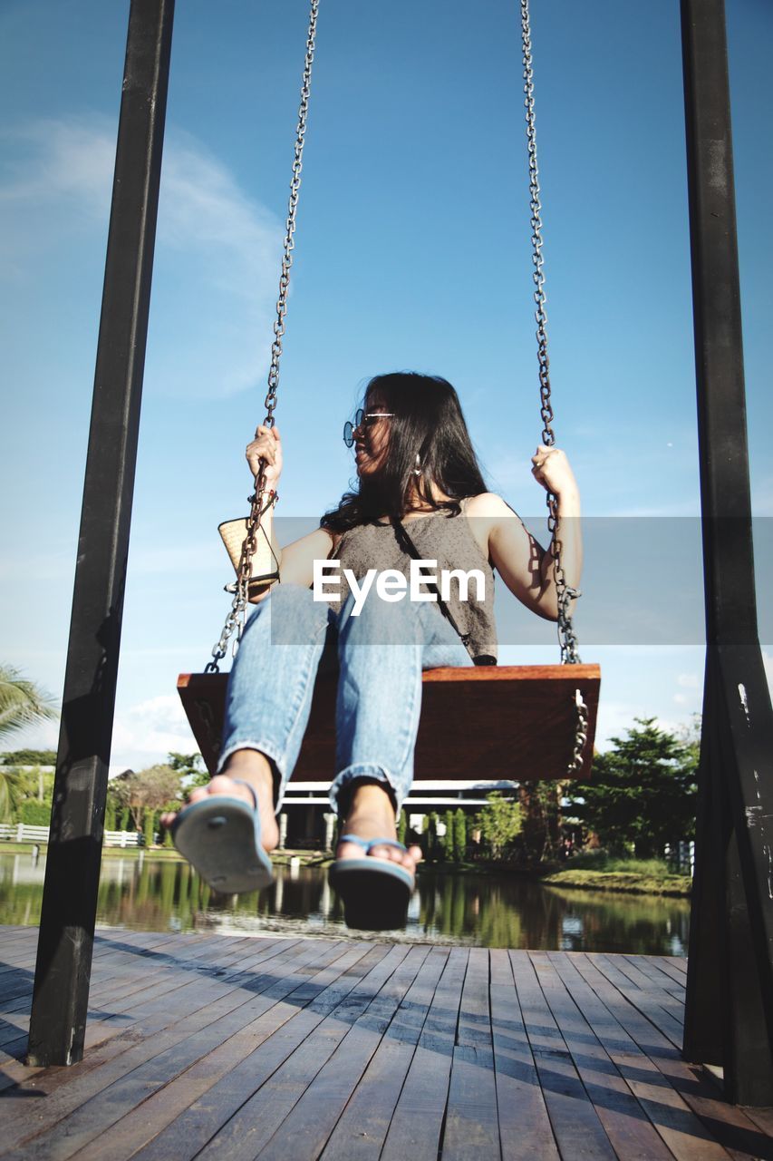 Woman sitting on swing at playground against lake and sky