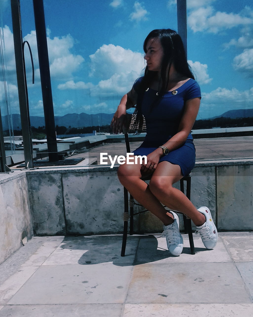 Young woman looking away while sitting on chair against blue sky during sunny day