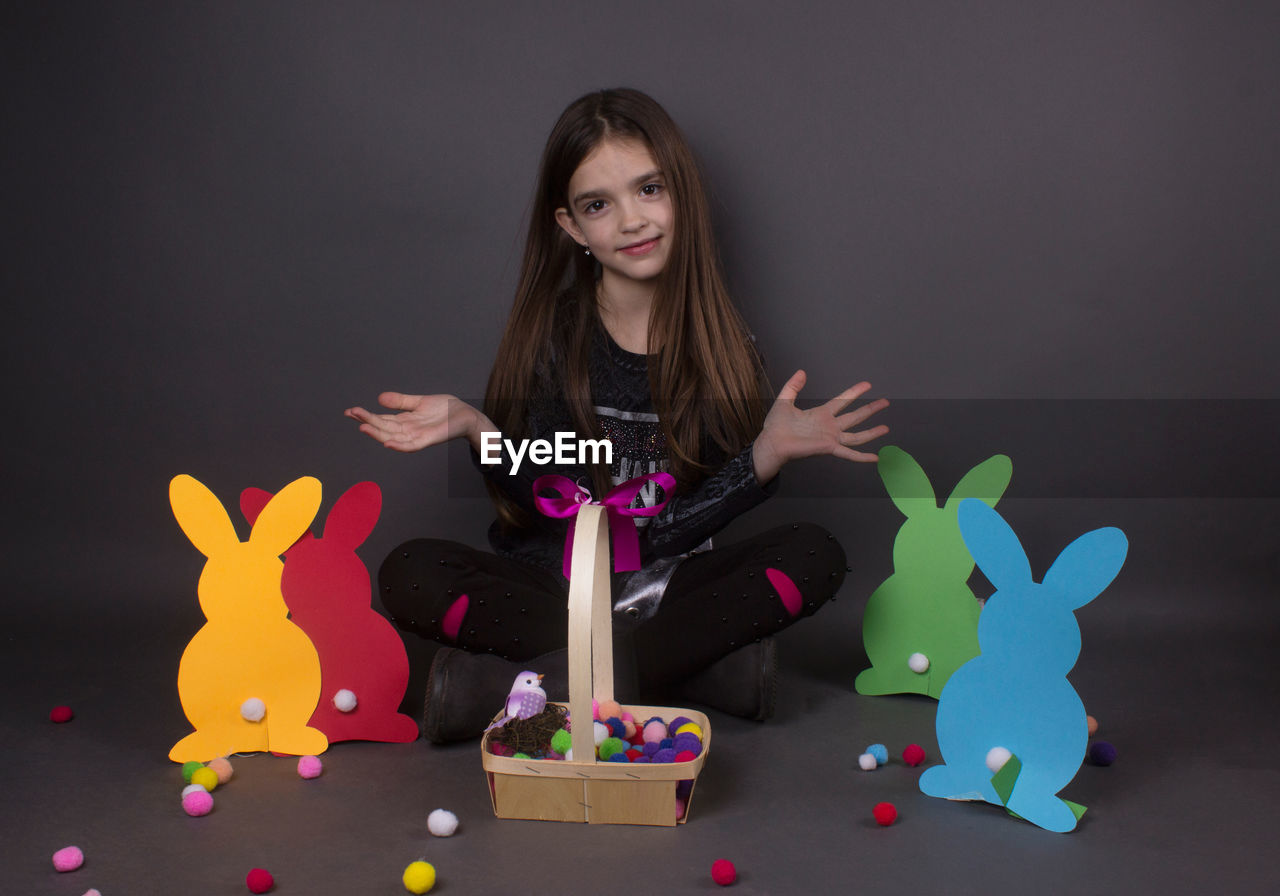 Portrait of smiling girl with eater bunny and pom pom in basket against gray background