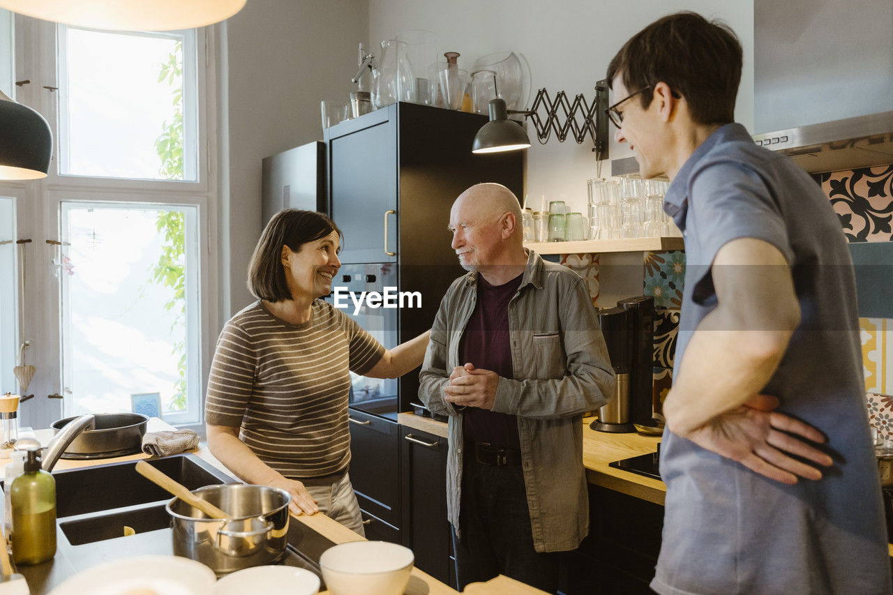 Smiling woman and man talking with senior father-in-law while standing in kitchen at home