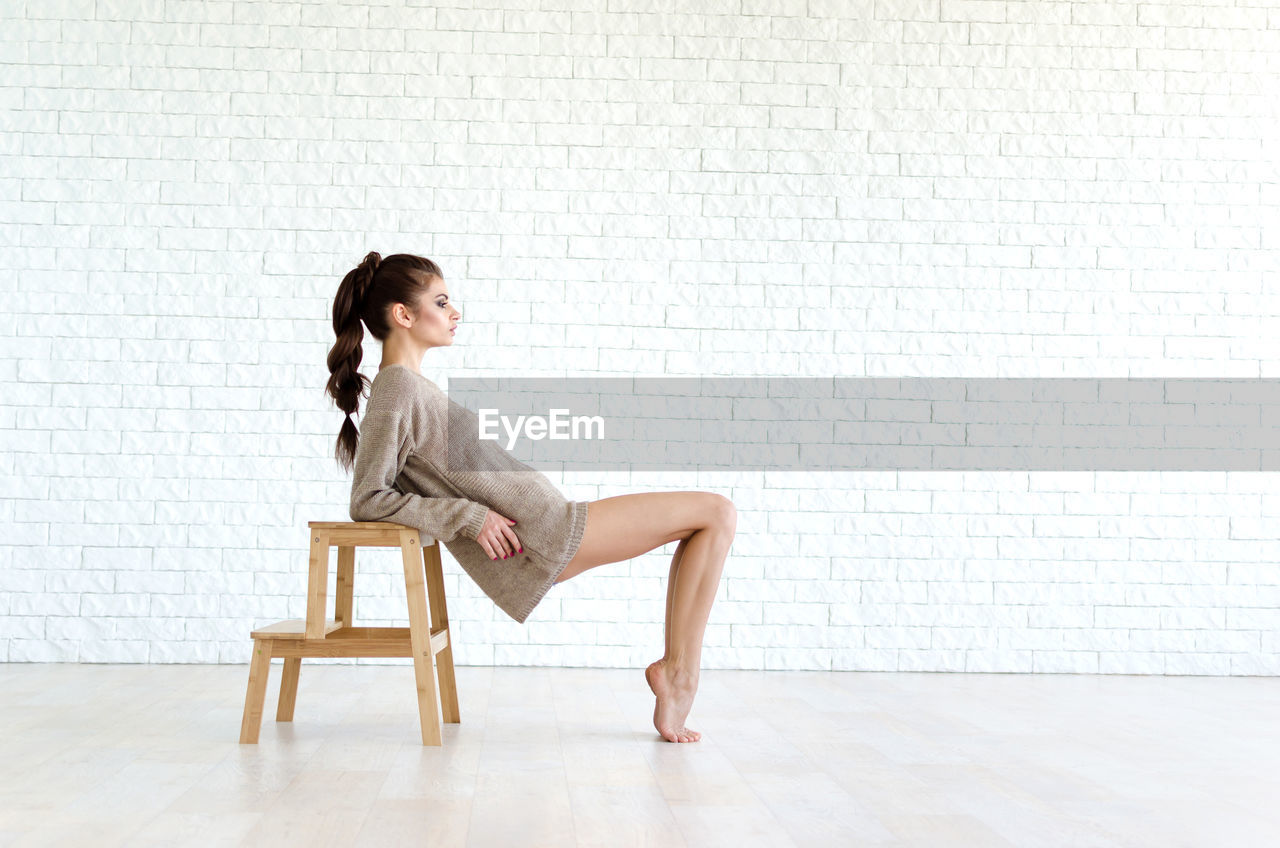 Full length of beautiful woman leaning on stool against brick wall
