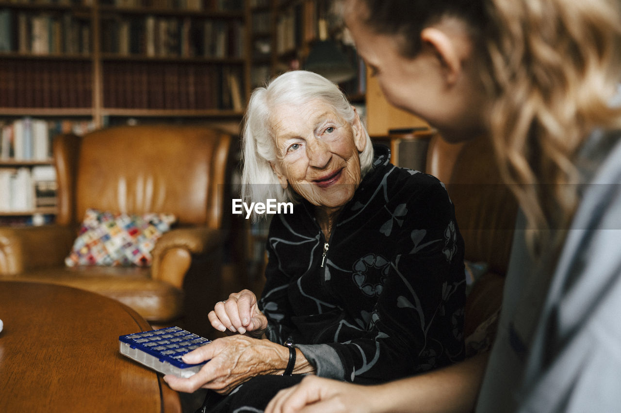 Smiling senior woman with medicine box talking with female healthcare worker in living room at home