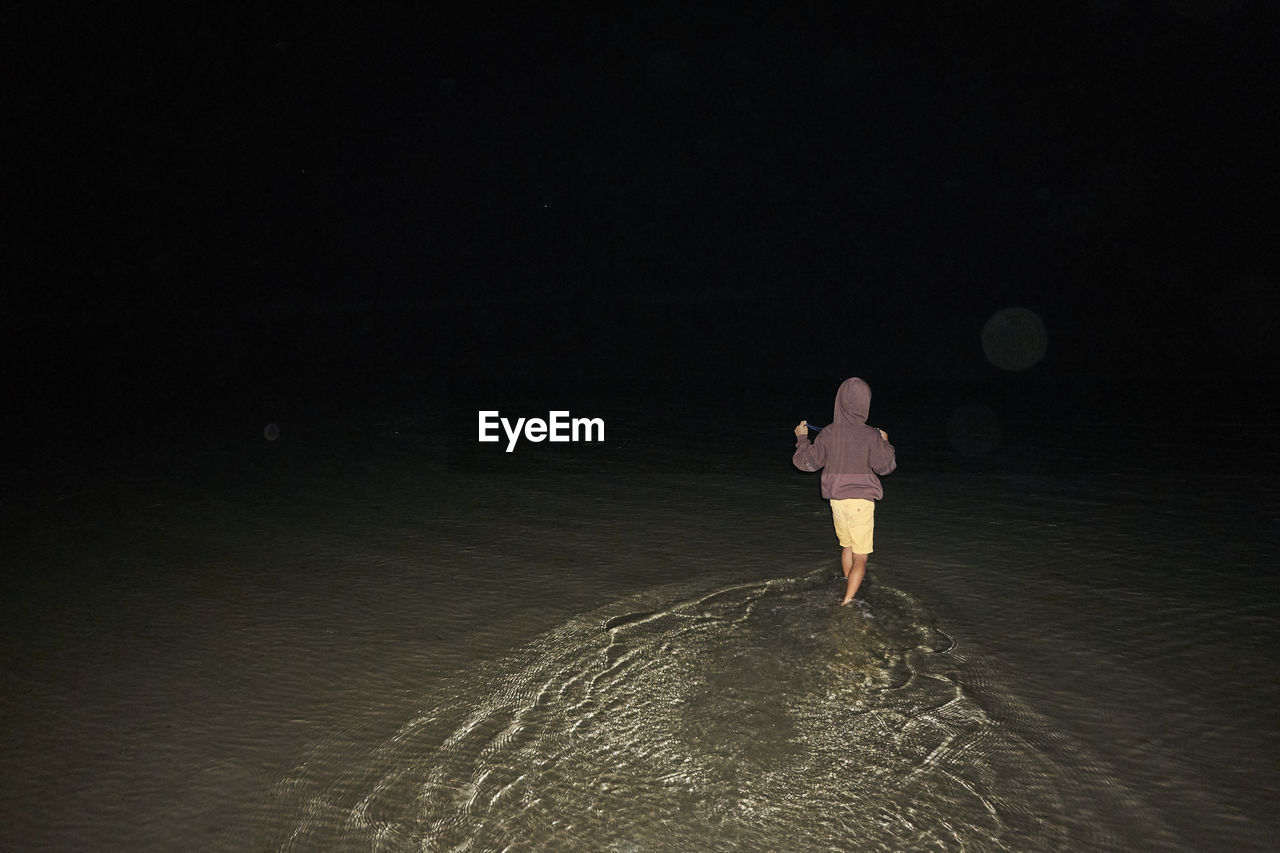 REAR VIEW OF WOMAN STANDING ON BEACH DURING NIGHT