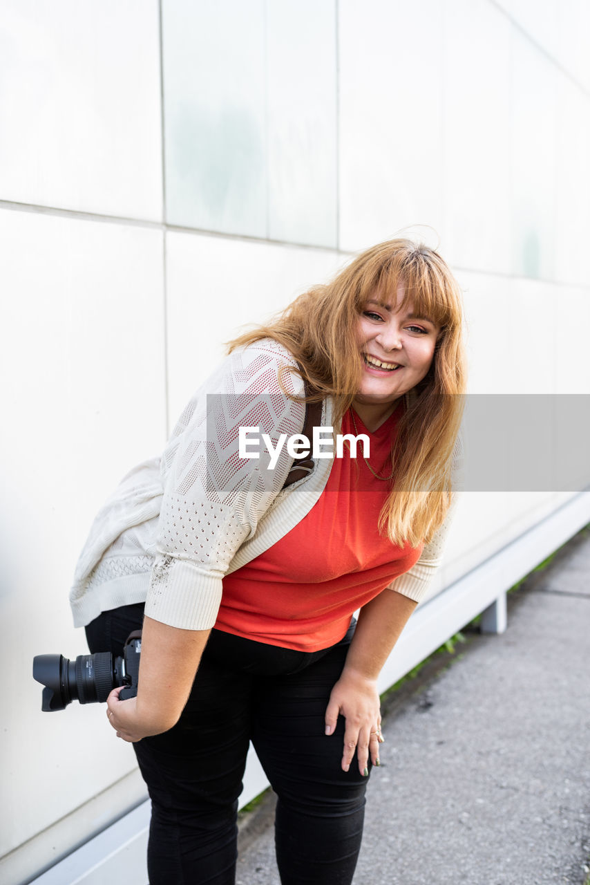 Portrait of smiling young woman holding camera standing against wall outdoors