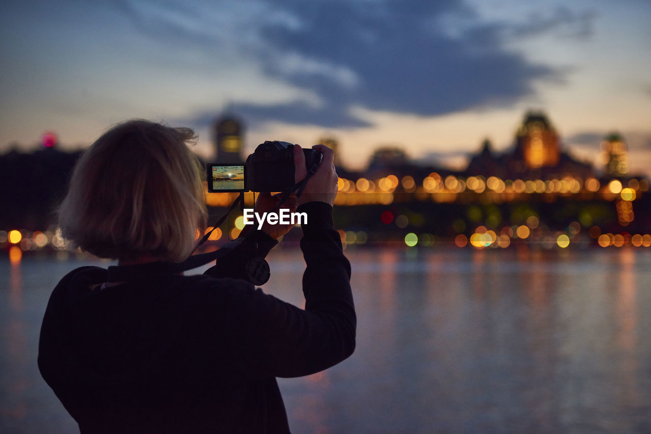 Rear view of woman photographing illuminated cityscape at sunset