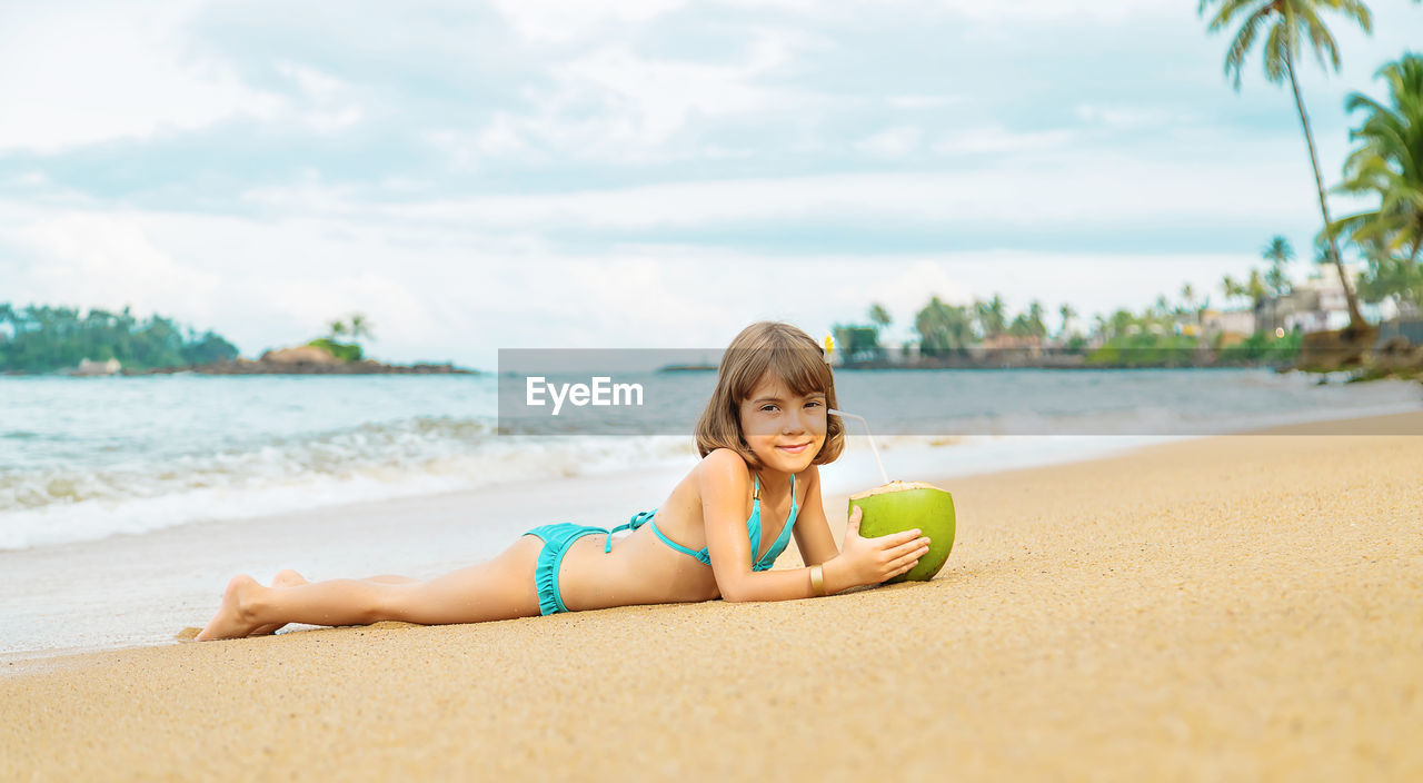 Portrait of smiling girl with coconut lying on beach