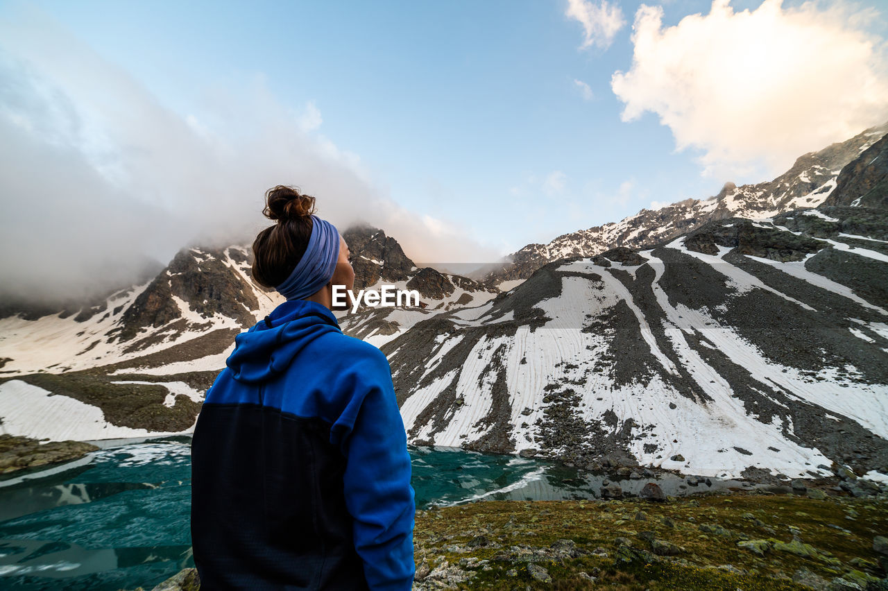 Woman stands near a frozen mountain lake, looking at the sky opening up from the fog and snow-capped