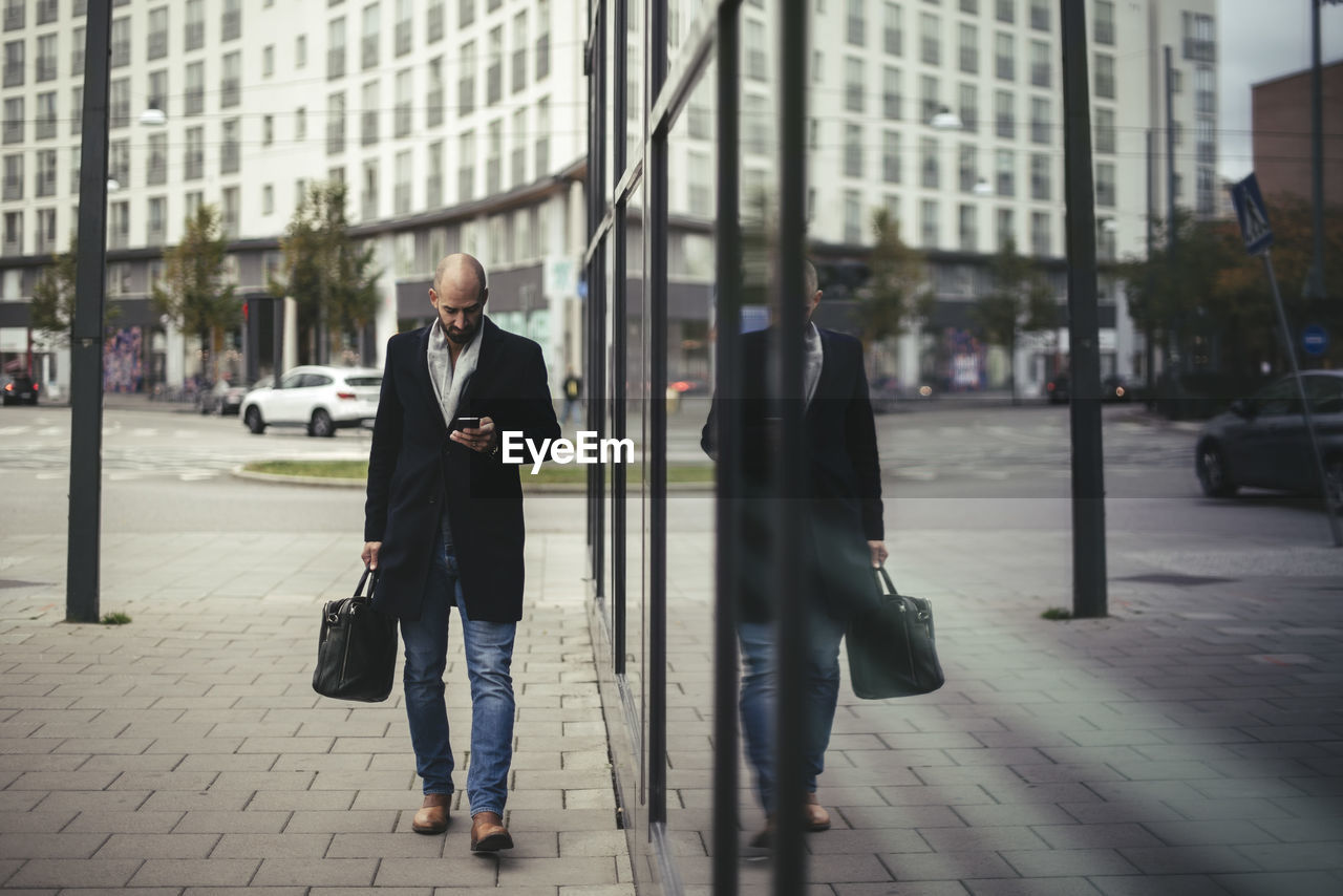 Full length of businessman using mobile phone while walking on sidewalk in city