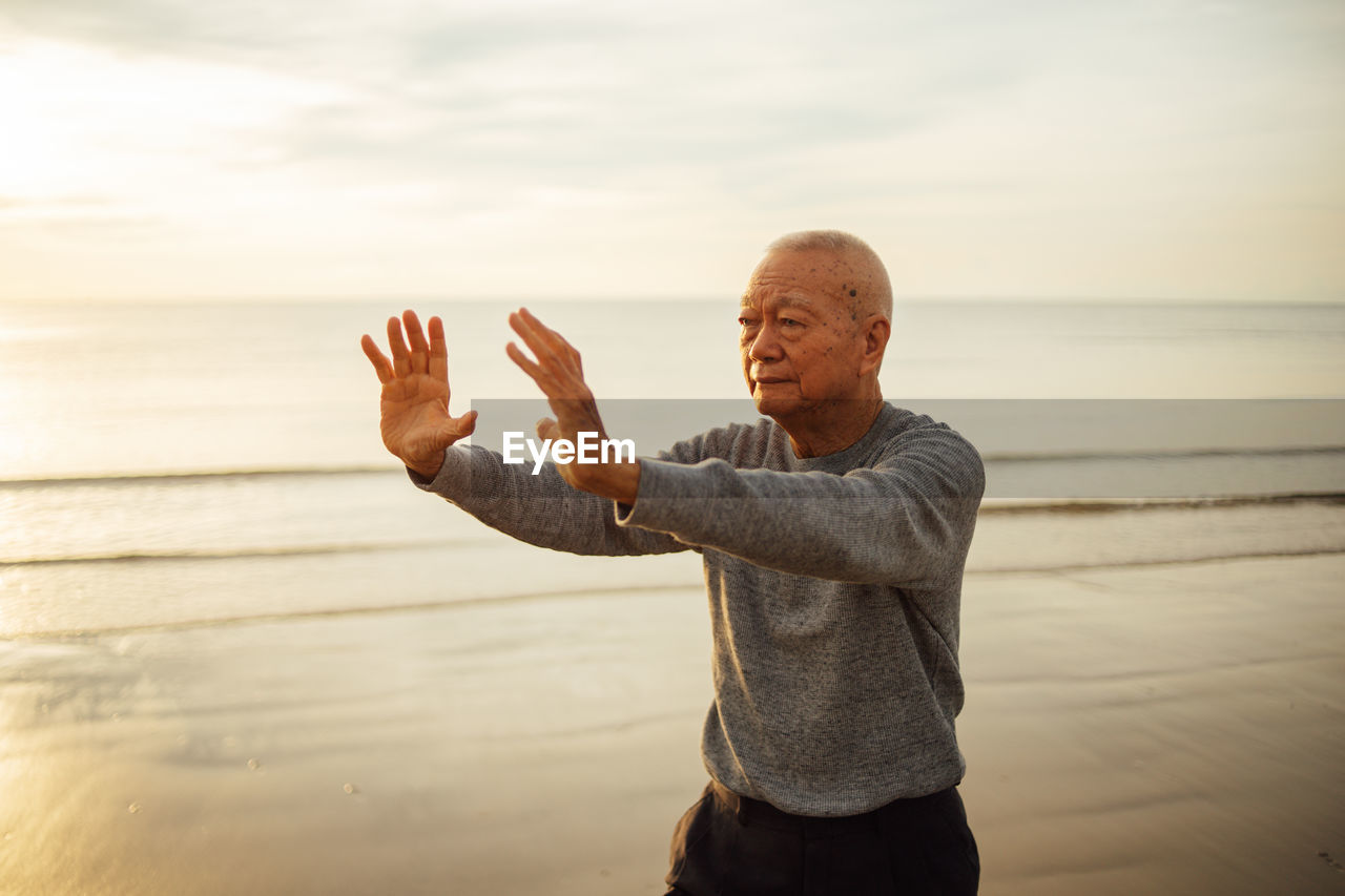 Senior man exercising while standing at beach against sky during sunset