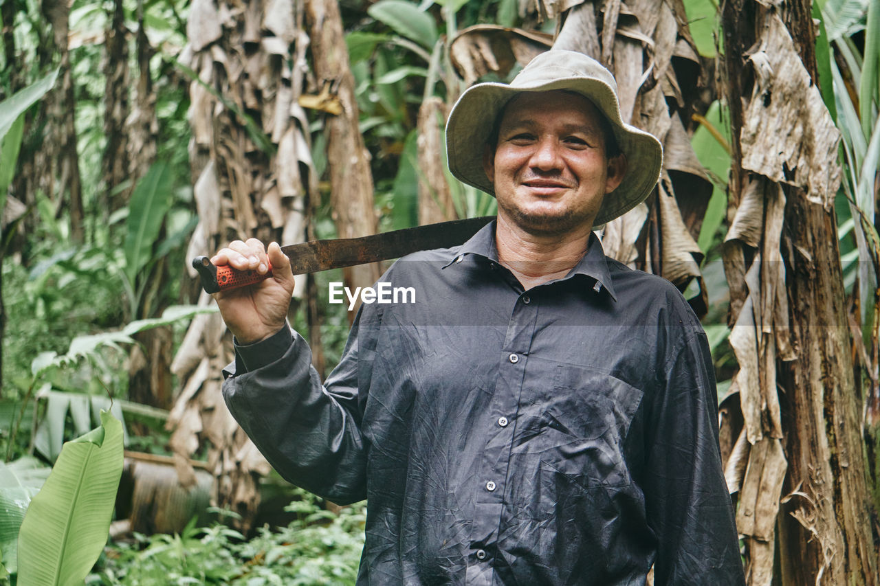 Positive hispanic man in shirt and bucket hat touching shoulder with machete and looking at camera with smile during work on ecological banana farm in costa rica