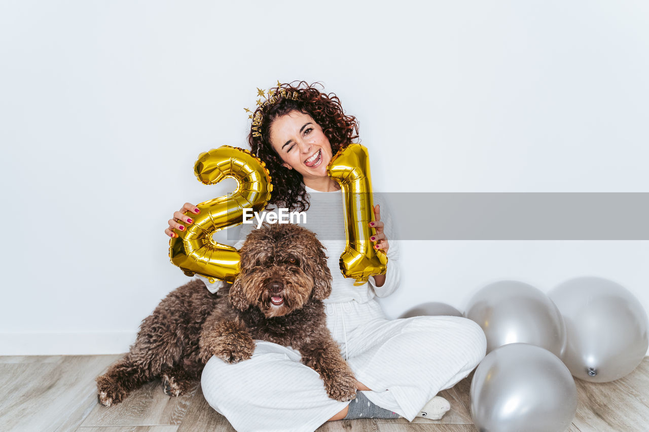 Portrait of smiling woman holding balloons sitting with dog against wall at home