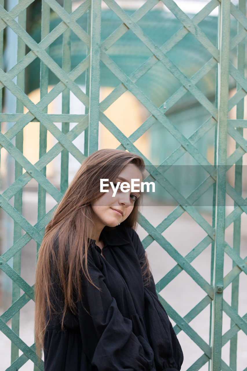 Portrait of young woman standing by metal grate outdoors