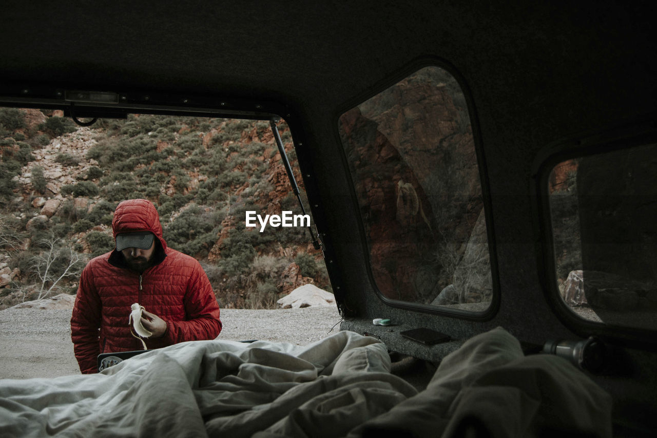 Man in warm clothing eating banana seen through off-road vehicle