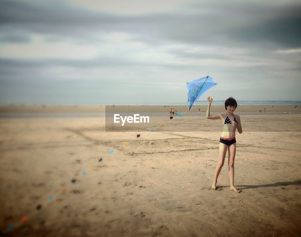 Full length of smiling teenage girl in bikini holding kite on beach against sky