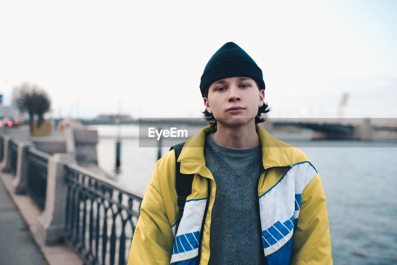 Handsome guy teenager boy 18-19 year old wear knit hat and trendy winter jacket posing at city view