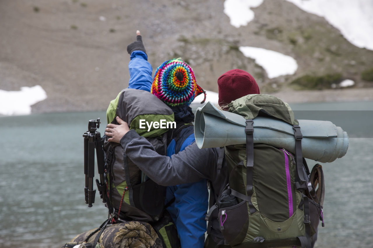 Rear view of hikers standing by lake against mountain during winter