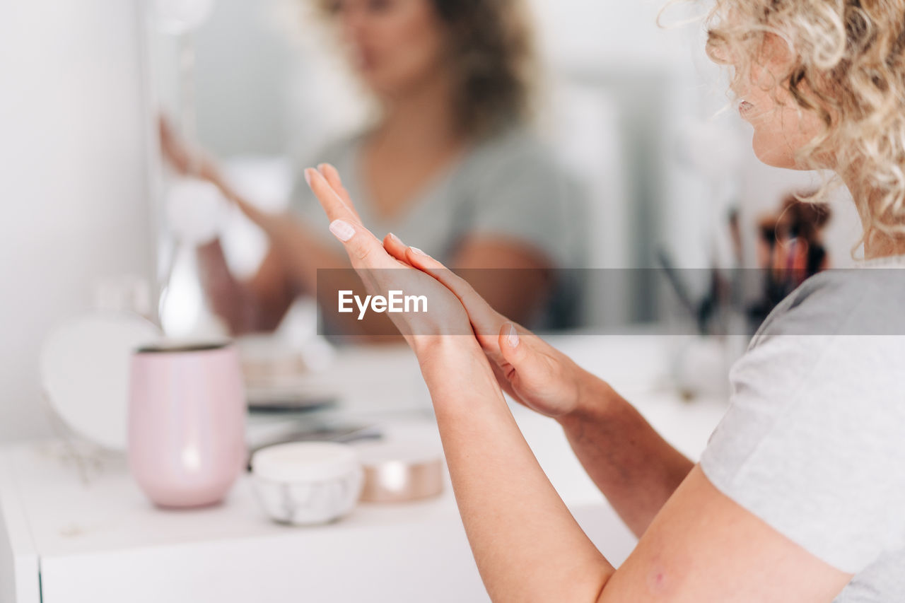 Crop female in casual shirt applying moisturizing hand cream while sitting at vanity table in light bedroom