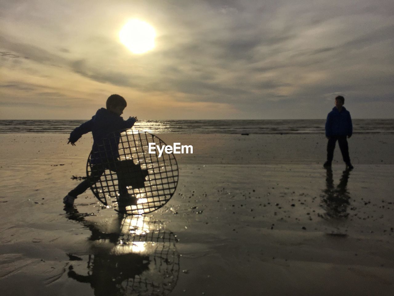 Boys playing at beach against sky during sunset