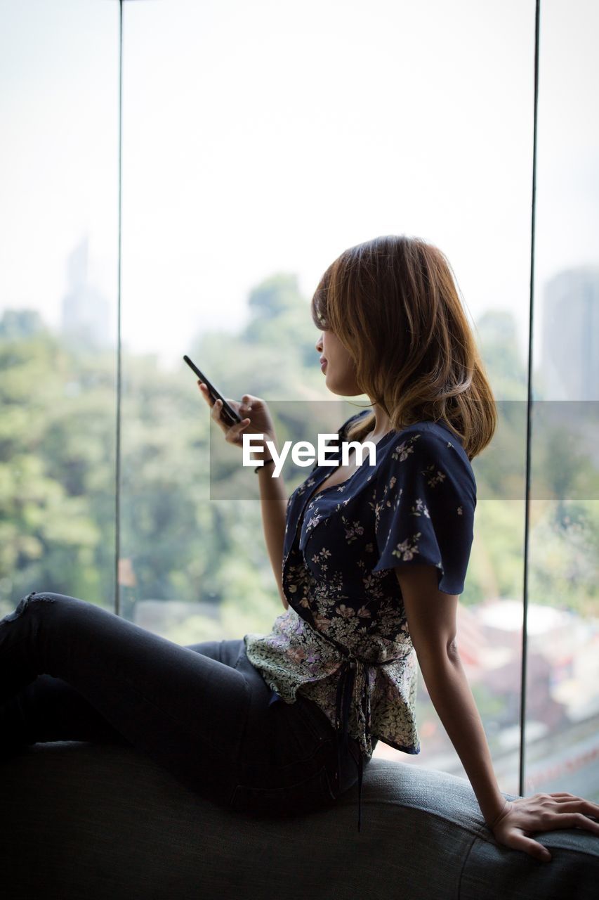 Side view of woman using phone while sitting by window