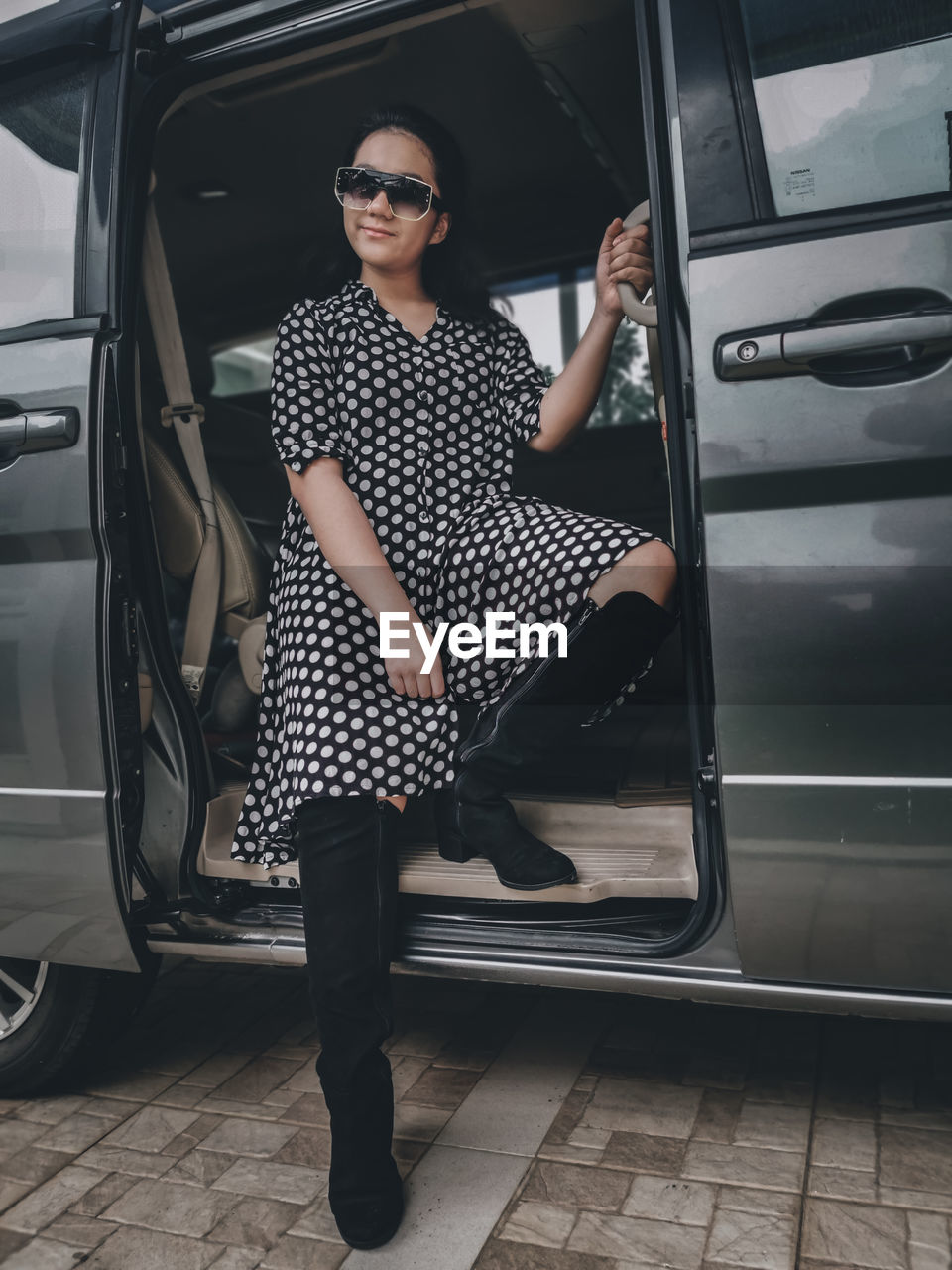 A girl wearing a polkadot dress, sun glasses and high boots sitting in a car 