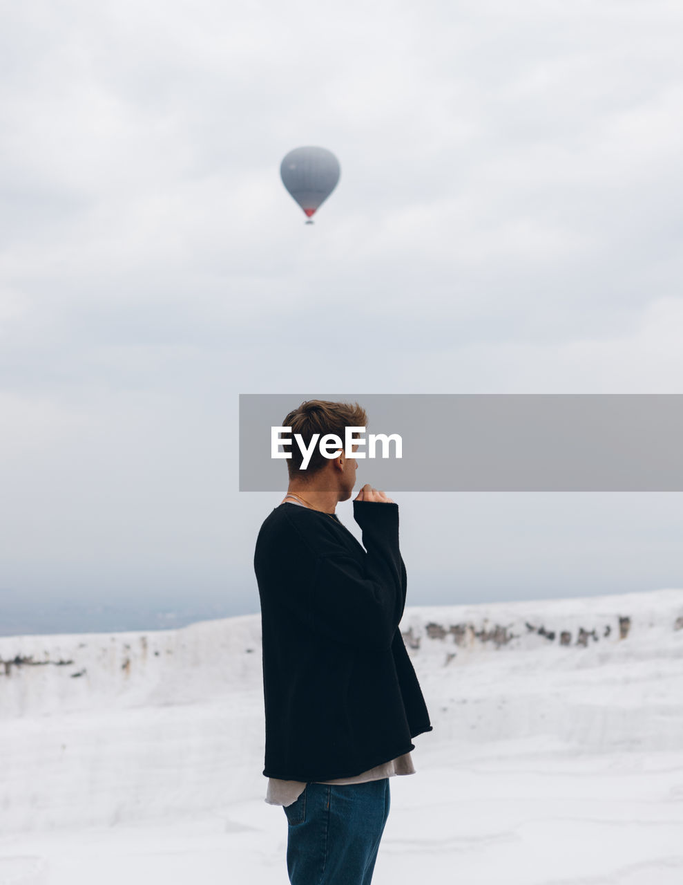 Traveling unrecognizable man in casual clothes looking away while standing on white hill of mineral formation against countryside at horizon and air balloon flying in gray sky in turkey