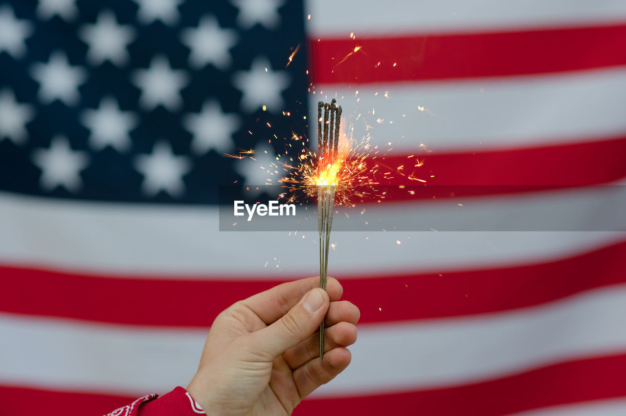 Cropped hand of person holding illuminated sparklers against american flag