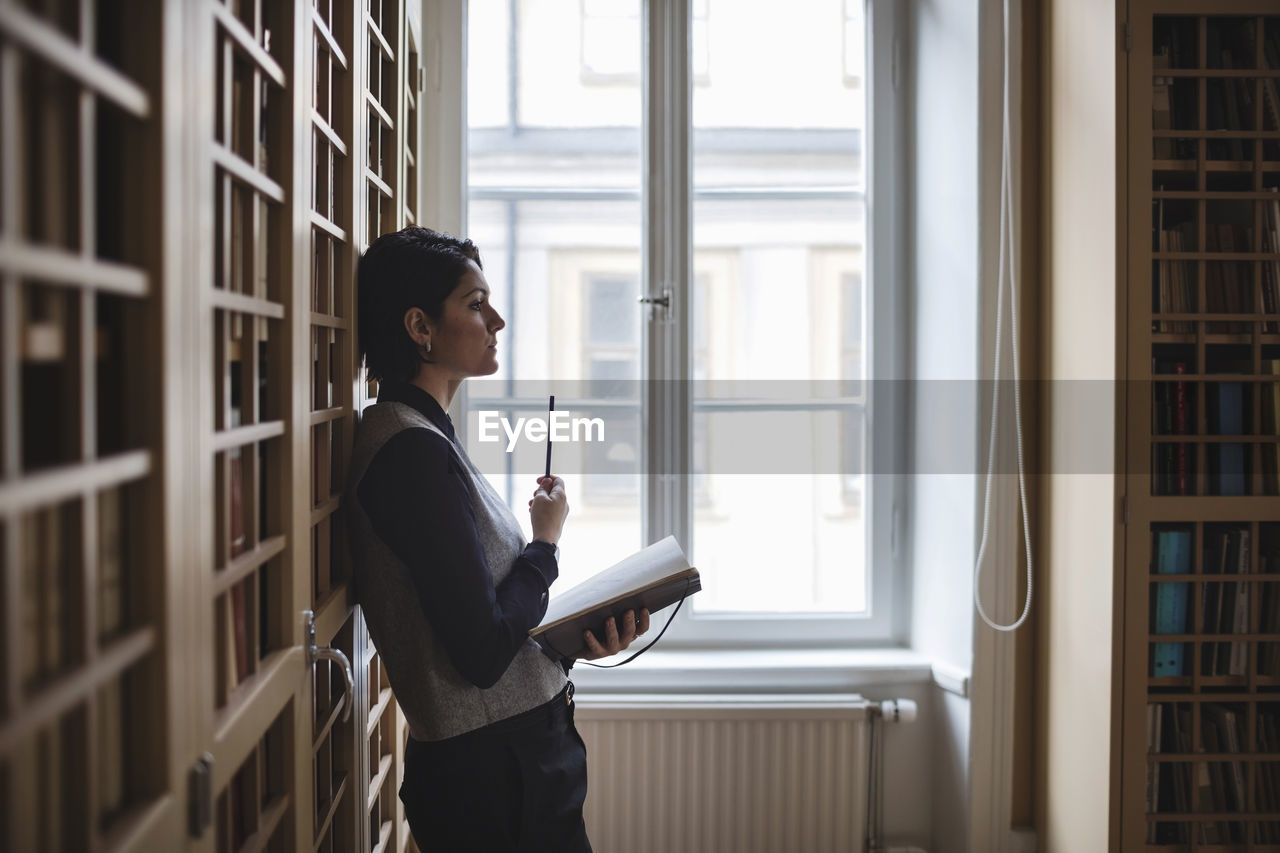 Profile view of thoughtful lawyer holding book while leaning on shelf in library