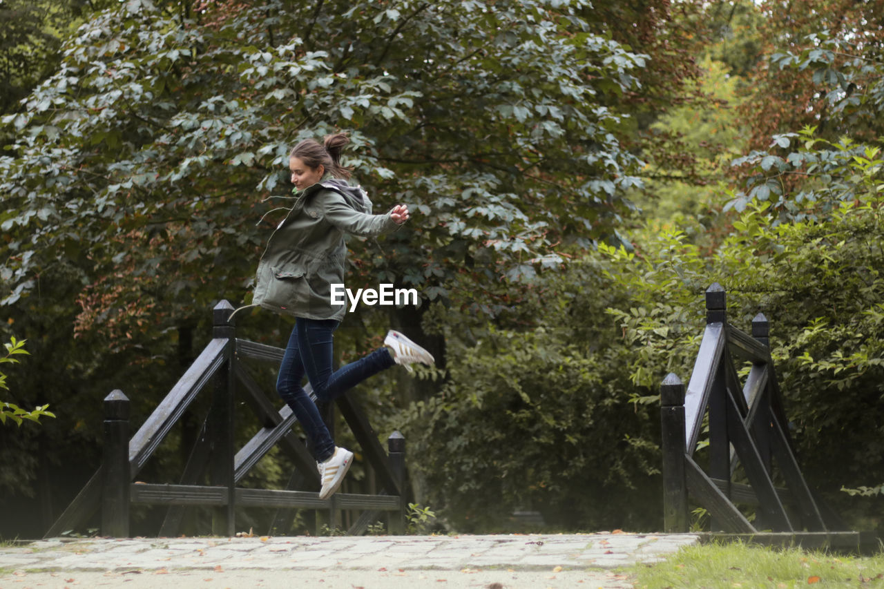 Side view of playful woman jumping on bridge against trees