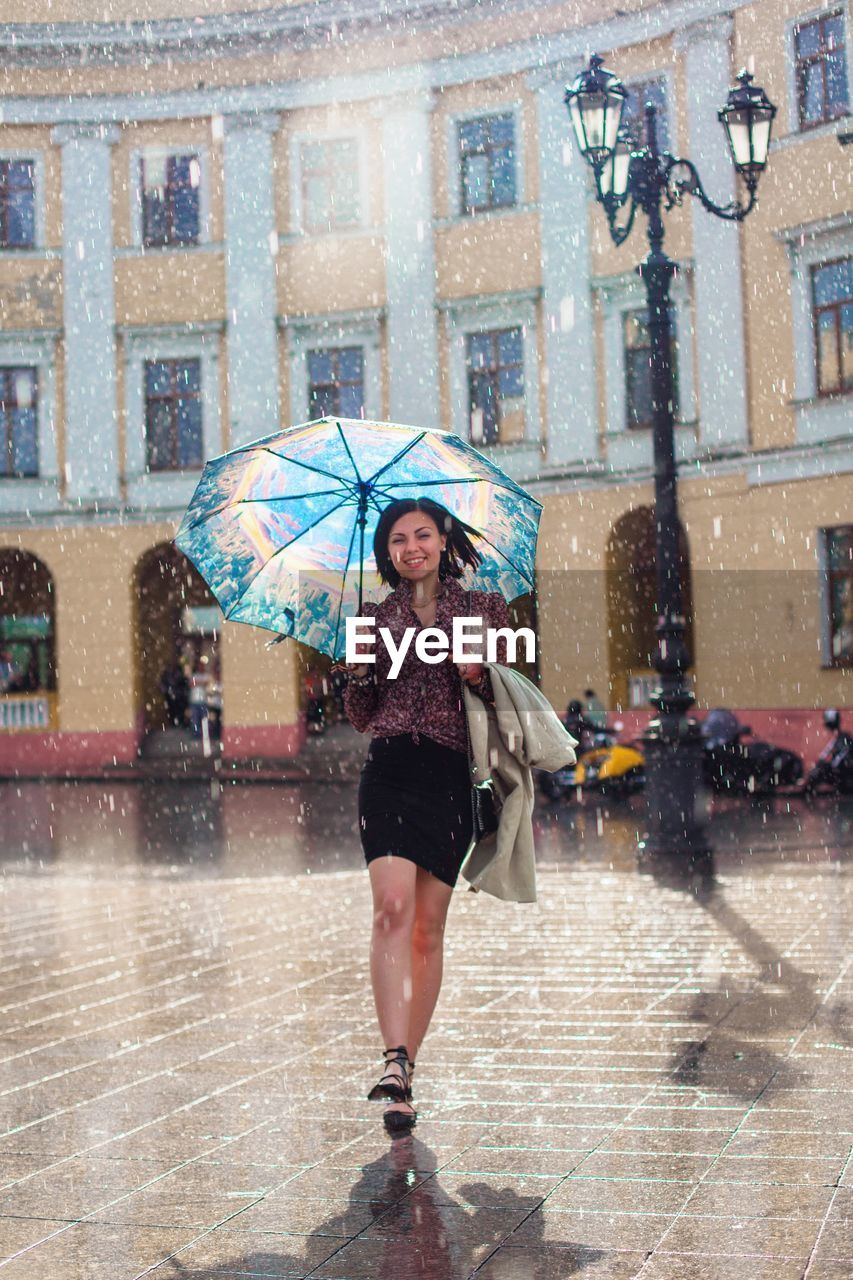 Full length portrait of cheerful woman walking with umbrella at fountain