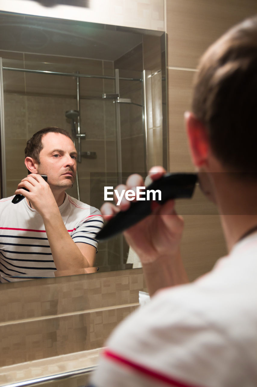 Young man reflecting on mirror while trimming beard in bathroom